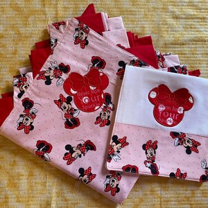 May include: Two pink and red fabric squares with a white fabric square in the middle. The pink fabric squares have a repeating pattern of Minnie Mouse with red polka dots. The white fabric square has a red embroidered Minnie Mouse with the text "Never lose your sparkle".