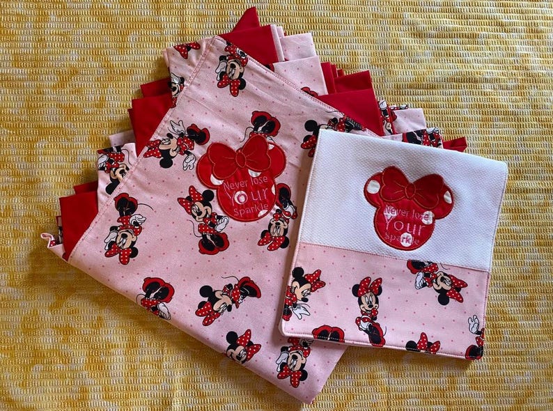 May include: Two pink and red fabric squares with a white fabric square in the middle. The pink fabric squares have a repeating pattern of Minnie Mouse with red polka dots. The white fabric square has a red embroidered Minnie Mouse with the text "Never lose your sparkle".