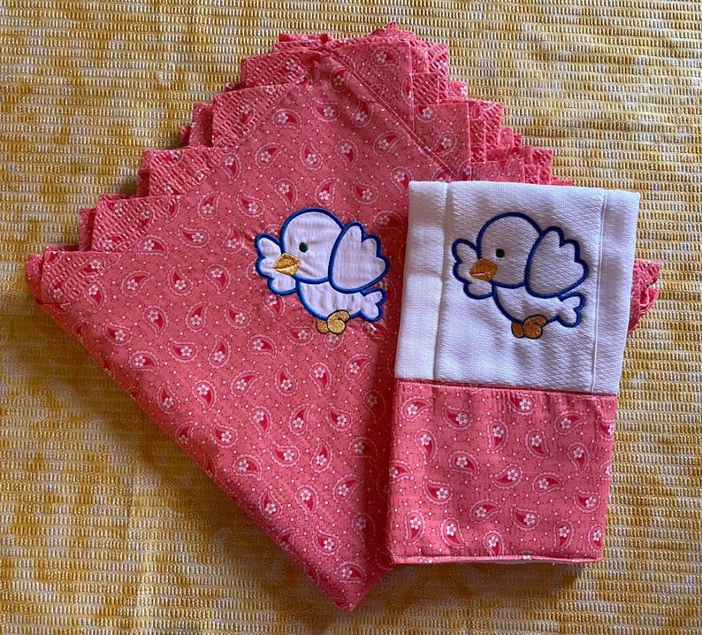 May include: A set of pink and white fabric napkins with embroidered birds. The napkins have a paisley pattern and are folded in a stack.