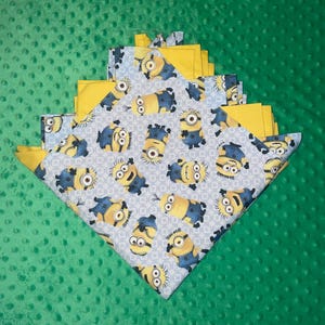 May include: A folded baby blanket with a blue and white pattern of Minions on one side and a solid yellow fabric on the other side. The blanket is folded in a triangular shape and is sitting on a green surface.