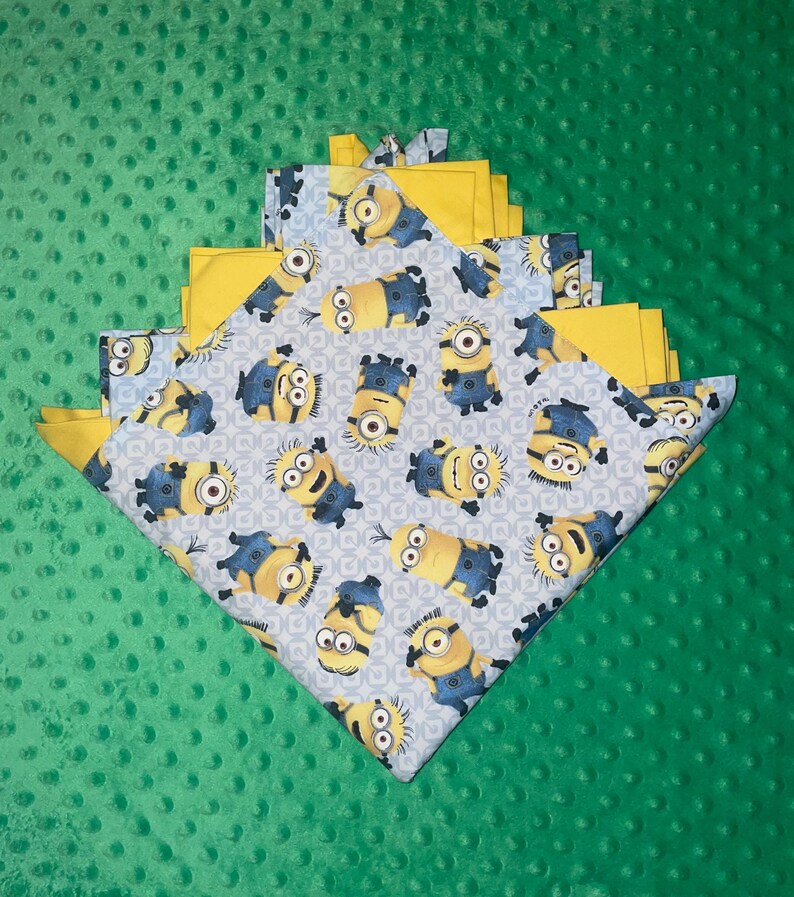 May include: A folded baby blanket with a blue and white pattern of Minions on one side and a solid yellow fabric on the other side. The blanket is folded in a triangular shape and is sitting on a green surface.
