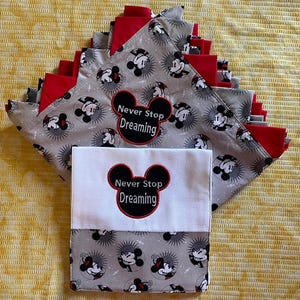 May include: A stack of gray and red fabric squares with a Mickey Mouse print. The squares have the text "Never Stop Dreaming" embroidered on them. The squares are folded in half and stacked on top of each other. The squares are on a yellow background.