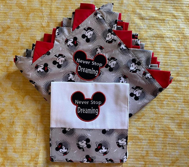 May include: A stack of gray and red fabric squares with a Mickey Mouse print. The squares have the text "Never Stop Dreaming" embroidered on them. The squares are folded in half and stacked on top of each other. The squares are on a yellow background.