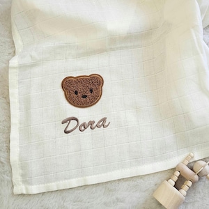 May include: A cream-coloured baby blanket with a grid pattern and a brown teddy bear applique. The name "Dora" is embroidered in brown script below the bear. A small wooden toy is in the bottom right corner.