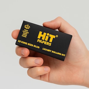 Puede incluir: Una caja rectangular negra de papel de liar "Hit Papers" se sostiene en una mano. La caja tiene texto dorado que dice "Hit Papers" y "Luxury Rolling Kit". La caja también tiene texto que dice "32 King Size Slim" y "100% Natural Wamp Ikoed Paper".