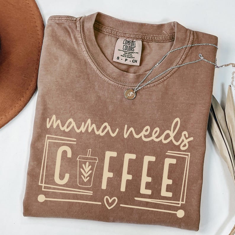 May include: A brown Comfort Colors t-shirt with the phrase "mama needs COFFEE" printed in a light beige color. The design includes a coffee cup graphic, a heart, and a rectangular border. A silver necklace with a pendant is draped across the shirt.
