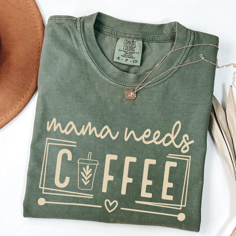 May include: An olive green t-shirt with the phrase "mama needs COFFEE" in a light tan color. The shirt features a coffee cup graphic and a small heart. A gold necklace is draped across the shirt.
