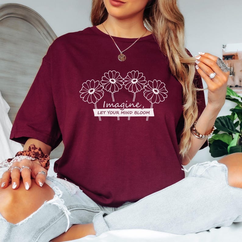 Pu&ograve; includere: Una t-shirt a maniche corte color bordeaux con una grafica a linee bianche raffigurante quattro margherite sul petto. Sotto i fiori, il testo "Imagine, LET YOUR MIND BLOOM" &egrave; scritto in una combinazione di corsivo e stampatello al centro.