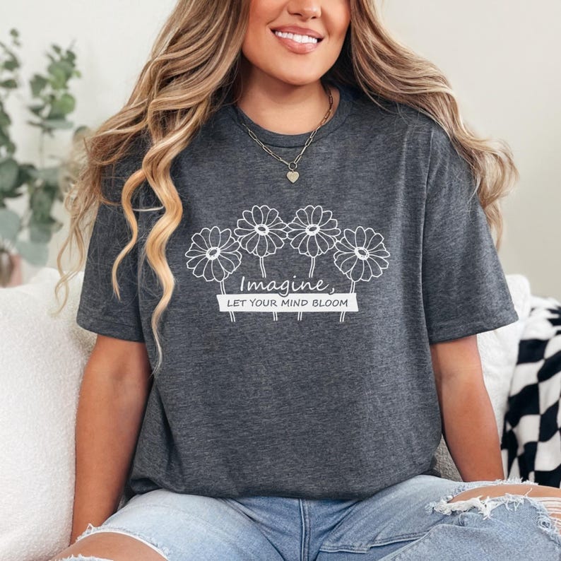 Pu&ograve; includere: Una t-shirt a maniche corte grigio scuro m&eacute;lange con una grafica a linee bianche raffigurante quattro fiori simili a margherite sopra il testo "Imagine, LET YOUR MIND BLOOM" stampato su un elemento rettangolare al centro del petto.