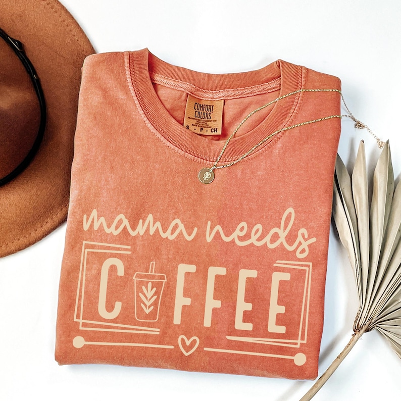 May include: Coral-colored Comfort Colors t-shirt with the phrase "mama needs COFFEE" in white. The design includes a coffee cup graphic, a heart, and a rectangular border. A gold necklace and a brown hat are also visible.