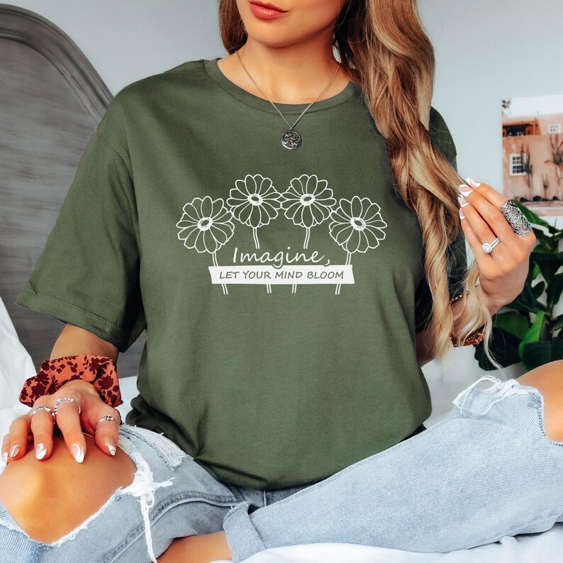 Pu&ograve; includere: Una t-shirt a maniche corte color verde oliva con una grafica a linee bianche di quattro margherite sopra il testo "Imagine, LET YOUR MIND BLOOM" stampato in un riquadro bianco. La maglietta ha un girocollo e una vestibilit&agrave; comoda.