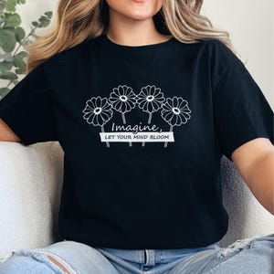 Pu&ograve; includere: Una t-shirt nera a maniche corte con una grafica a linee bianche che raffigura quattro fiori simili a margherite. Sotto i fiori, il testo "Imagine, LET YOUR MIND BLOOM" &egrave; scritto in una combinazione di corsivo e stampatello.
