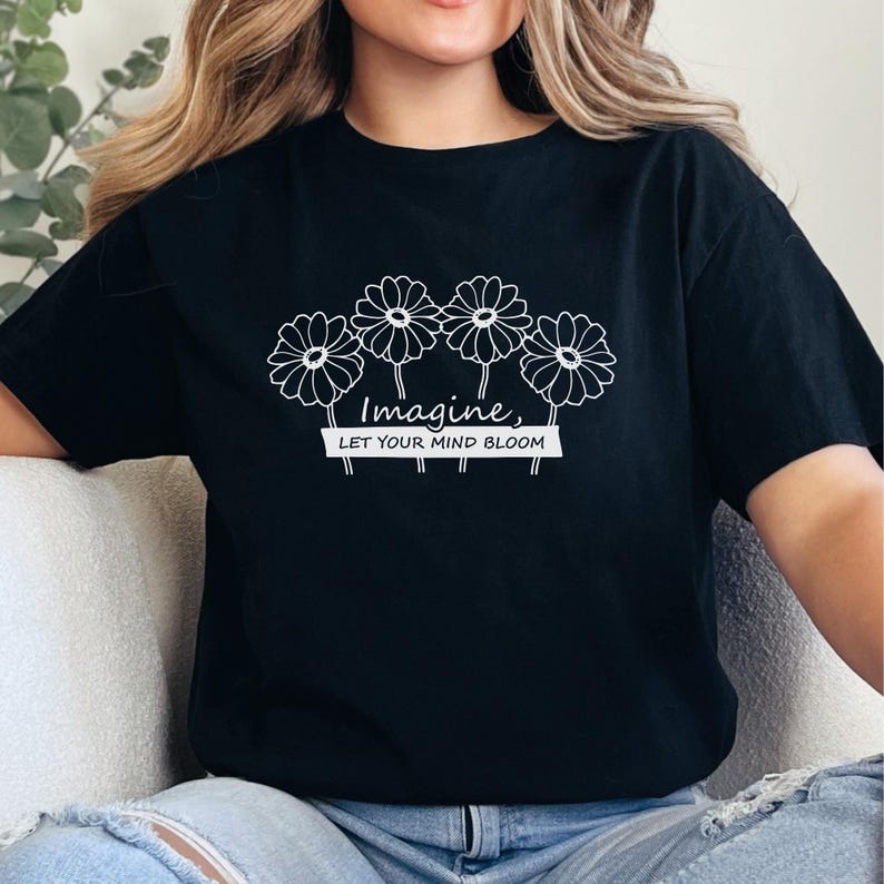 Pu&ograve; includere: Una t-shirt nera a maniche corte con una grafica a linee bianche che raffigura quattro fiori simili a margherite. Sotto i fiori, il testo "Imagine, LET YOUR MIND BLOOM" &egrave; scritto in una combinazione di corsivo e stampatello.