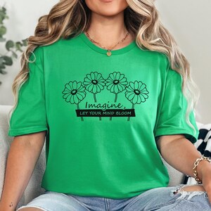Pu&ograve; includere: Una t-shirt a maniche corte di colore verde brillante con una grafica a linee nere raffigurante quattro margherite. Sotto i fiori, il testo "Imagine, LET YOUR MIND BLOOM" &egrave; scritto in una combinazione di corsivo e stampatello sul petto.