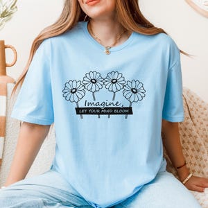 Pu&ograve; includere: Una t-shirt a maniche corte color azzurro con una grafica a linee nere raffigurante quattro margherite. Sotto i fiori, il testo "Imagine, LET YOUR MIND BLOOM" &egrave; stampato in una combinazione di corsivo e stampatello sul petto.
