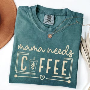 May include: A teal Comfort Colors t-shirt with the phrase "mama needs COFFEE" in a cream-colored font. The design includes a coffee cup graphic and a heart. A gold necklace with a pendant is draped across the shirt.
