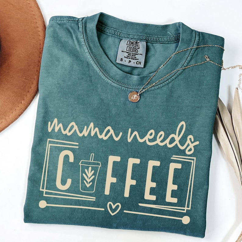 May include: A teal Comfort Colors t-shirt with the phrase "mama needs COFFEE" in a cream-colored font. The design includes a coffee cup graphic and a heart. A gold necklace with a pendant is draped across the shirt.