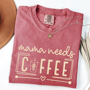 May include: A red-toned t-shirt with the phrase "mama needs COFFEE" in a light beige color. The design includes a coffee cup graphic and a heart. A silver necklace with a pendant is draped across the shirt.