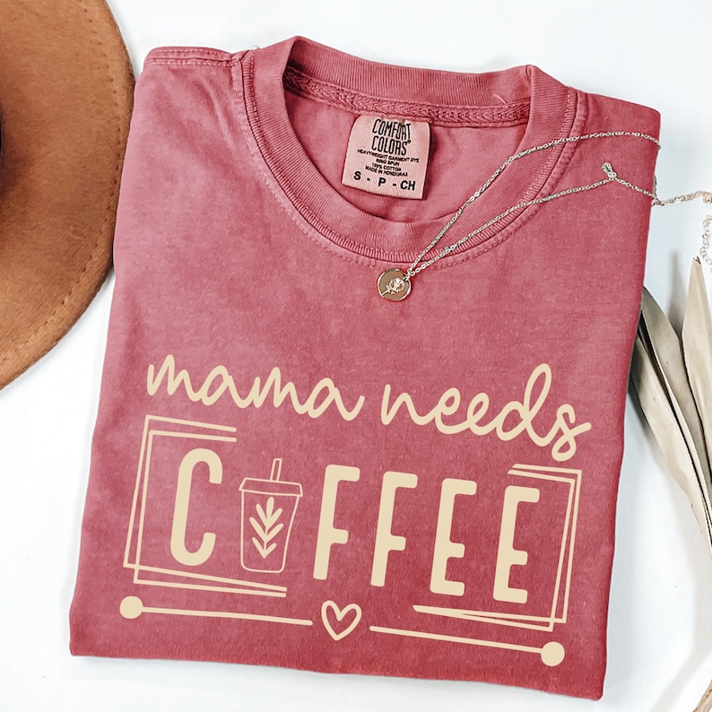 May include: A red-toned t-shirt with the phrase "mama needs COFFEE" in a light beige color. The design includes a coffee cup graphic and a heart. A silver necklace with a pendant is draped across the shirt.
