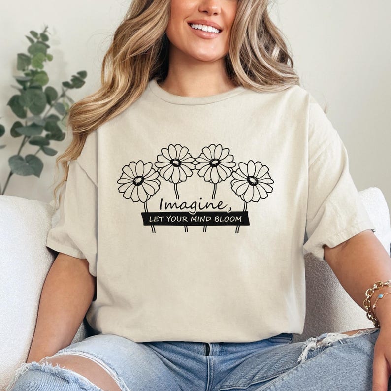 Pu&ograve; includere: Una t-shirt beige a maniche corte con una grafica a linee nere raffigurante quattro fiori simili a margherite. Sotto i fiori, il testo "Imagine, LET YOUR MIND BLOOM" &egrave; stampato in una combinazione di corsivo e stampatello sul petto.