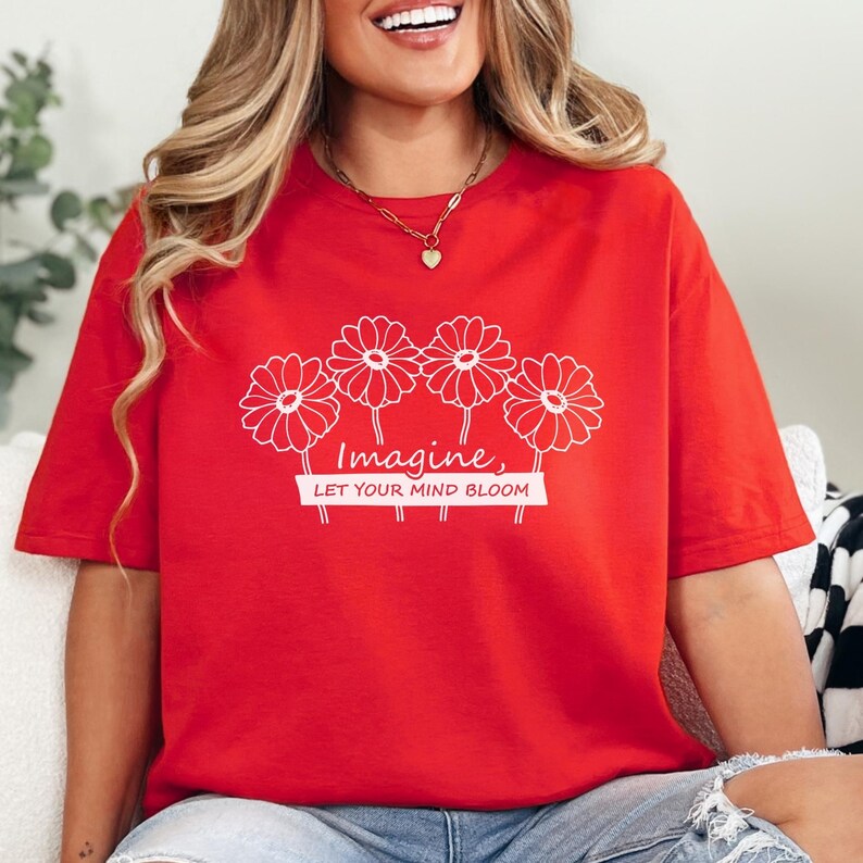 Pu&ograve; includere: Una t-shirt rossa a maniche corte con una grafica a linee bianche raffigurante quattro fiori simili a margherite. Sotto i fiori, il testo "Imagine, LET YOUR MIND BLOOM" &egrave; scritto in una combinazione di caratteri corsivi e sans-serif.