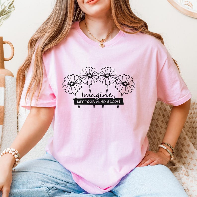 Pu&ograve; includere: Una t-shirt a maniche corte color rosa chiaro con una grafica nera di quattro fiori simili a margherite sopra il testo "Imagine, LET YOUR MIND BLOOM" stampato su uno sfondo rettangolare nero. La maglietta ha un girocollo.
