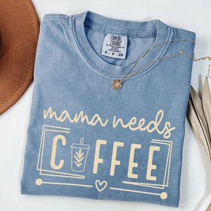 May include: A blue Comfort Colors t-shirt with the phrase "mama needs COFFEE" in a light tan color. The design includes a coffee cup graphic and a heart. A gold necklace with a pendant is draped across the shirt.