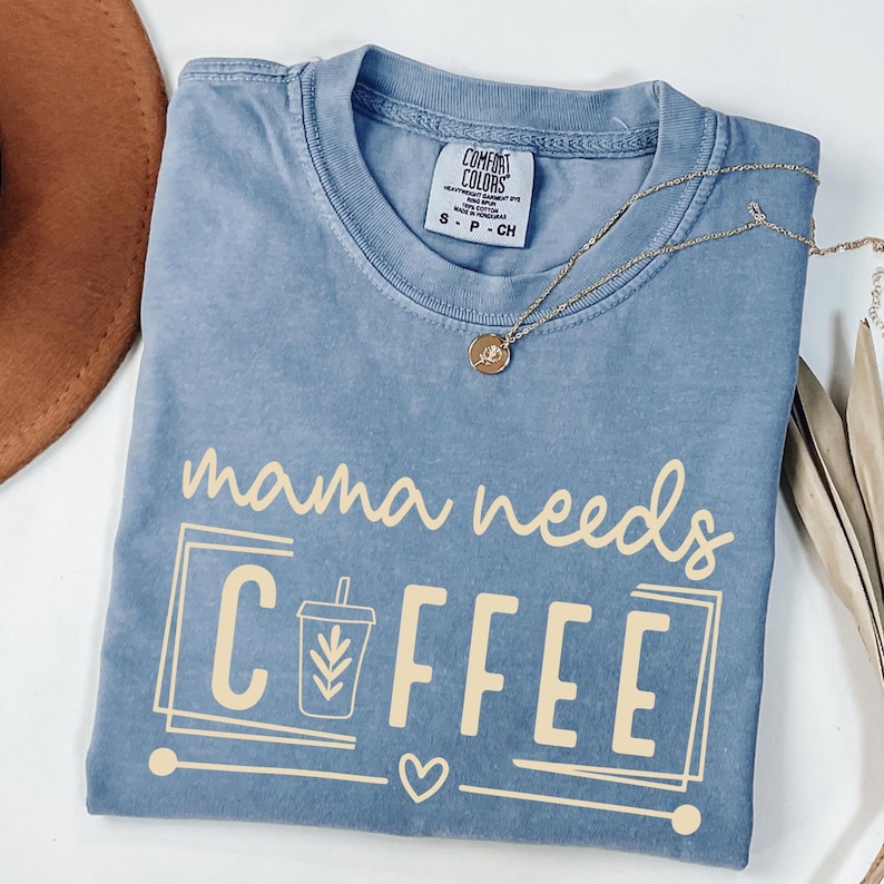 May include: A blue Comfort Colors t-shirt with the phrase "mama needs COFFEE" in a light tan color. The design includes a coffee cup graphic and a heart. A gold necklace with a pendant is draped across the shirt.