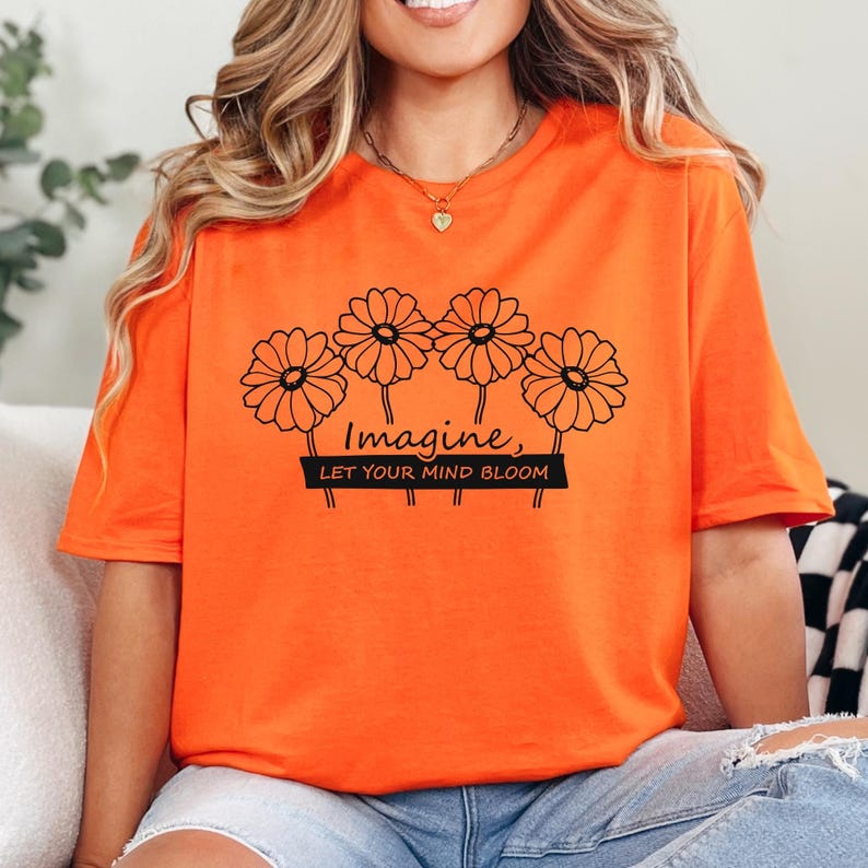 Pu&ograve; includere: Una t-shirt a maniche corte color arancione brillante con un disegno a linee nere di quattro margherite. Sotto i fiori, il testo "Imagine, LET YOUR MIND BLOOM" &egrave; scritto in nero, con la seconda riga su una barra rettangolare nera.