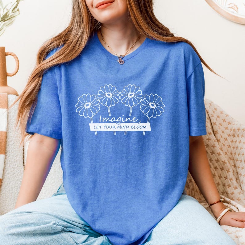 Pu&ograve; includere: Una t-shirt blu brillante a maniche corte con una grafica a linee bianche di quattro margherite sopra il testo "Imagine, LET YOUR MIND BLOOM" stampato in un carattere sans-serif pulito. La maglietta ha un classico girocollo.