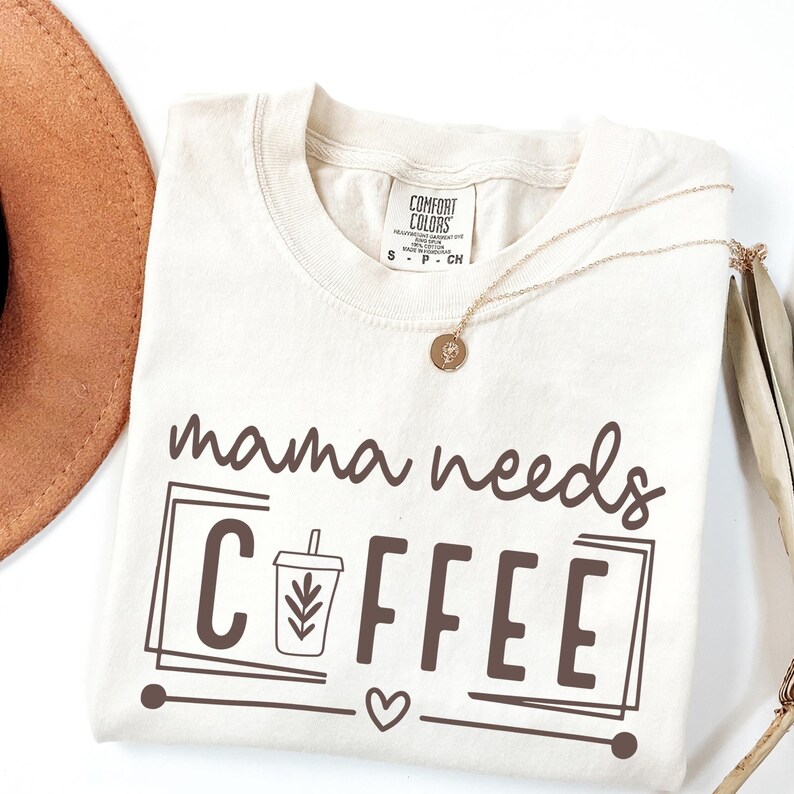 May include: Cream-colored t-shirt with the phrase "mama needs COFFEE" in brown lettering. The design includes a coffee cup graphic and a heart. A gold necklace and a brown hat are partially visible.