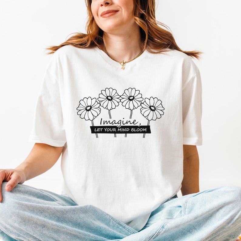 Pu&ograve; includere: Una t-shirt bianca a maniche corte con una grafica a linee nere di quattro margherite sopra il testo "Imagine, LET YOUR MIND BLOOM" stampato su uno sfondo rettangolare nero. La maglietta presenta un girocollo e una vestibilit&agrave; ampia.