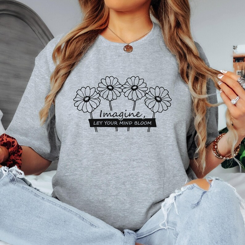 Pu&ograve; includere: Una t-shirt grigio melange a girocollo con una grafica a linee nere raffigurante quattro margherite disposte in fila. Sotto i fiori, il testo "Imagine, LET YOUR MIND BLOOM" &egrave; scritto in una combinazione di caratteri corsivi e sans-serif.