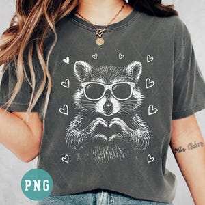 May include: Dark gray t-shirt featuring a white graphic of a raccoon wearing sunglasses and forming a heart with its hands. White hearts surround the raccoon. The shirt is a casual, unisex style.