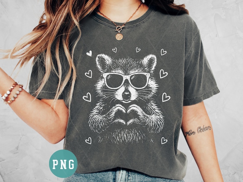 May include: Dark gray t-shirt featuring a white graphic of a raccoon wearing sunglasses and forming a heart with its hands. White hearts surround the raccoon. The shirt is a casual, unisex style.