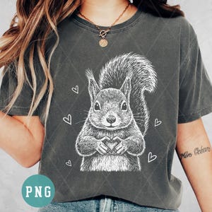 Op de afbeelding: Donkergrijs T-shirt met een gedetailleerde witte illustratie van een eekhoorn die met zijn handen een hart vormt. Witte harten zijn verspreid rond de eekhoorn. Het shirt heeft een ruitpatroon.