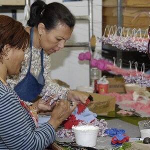 May include: Two people are working on a craft project. One person is holding a blue and white flower and the other person is holding a red and white flower. There are many other flowers in the background.