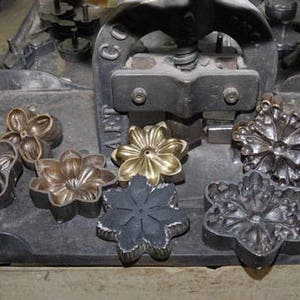 May include: A close-up of a metal press with several metal flower-shaped molds. The molds are arranged on a black metal surface. The press is used to create decorative metal flowers.