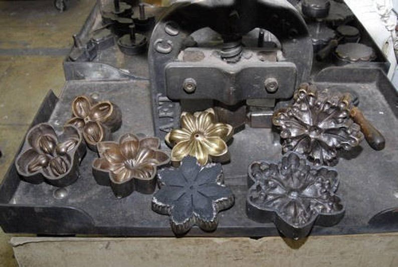 May include: A close-up of a metal press with several metal flower-shaped molds. The molds are arranged on a black metal surface. The press is used to create decorative metal flowers.