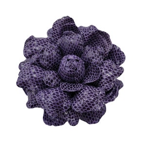 May include: A purple flower brooch with a textured, scaly surface. The flower is made of fabric and has a realistic appearance.