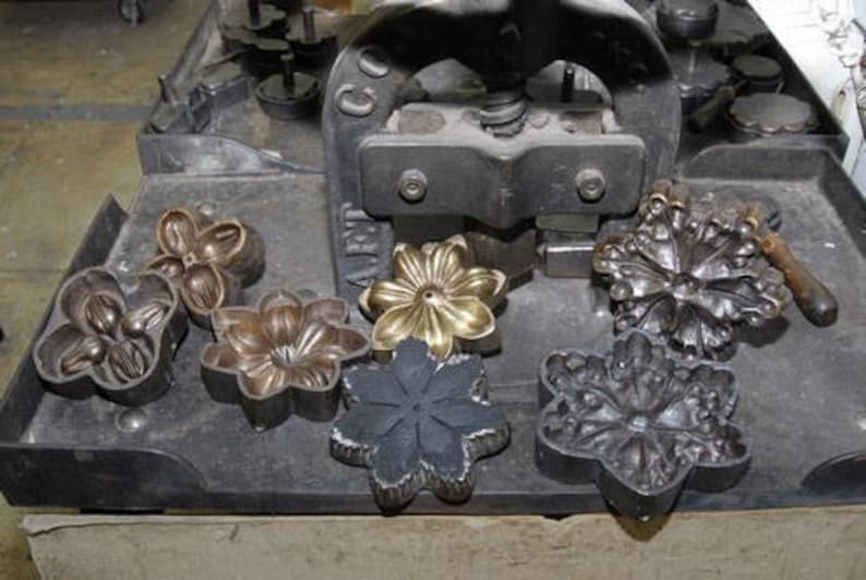 May include: A close-up of a metal press with several different metal flower-shaped molds. The molds are arranged on a black metal surface. The press is used to create decorative metal flowers.