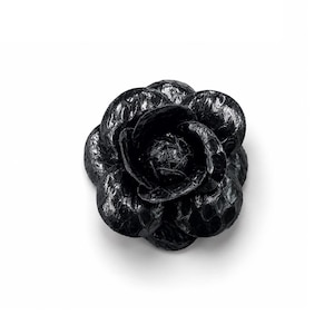 May include: A black flower-shaped brooch featuring a textured, scale-like surface pattern. The accessory is constructed with layered, rounded petals that spiral toward a central point, creating a dimensional and realistic floral appearance.