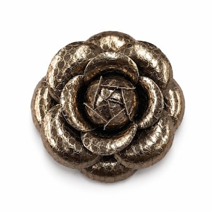 May include: A bronze-colored flower brooch with a metallic finish and a textured, scale-like pattern on the petals. The accessory is shaped like a camellia with multiple overlapping layers of rounded petals arranged in a circular, blooming design.