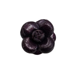 Könnte beinhalten: Eine dunkelviolette Blumenbrosche mit einem strukturierten, schuppenartigen Muster auf den abgerundeten Blütenblättern. Das Accessoire ist wie eine Kamelie geformt, mit geschichteten, überlappenden Blütenblättern, die eine dichte, kreisförmige Blüte mit einer zentralen Knospe bilden.