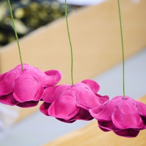 May include: A string of pink paper flowers hanging from green strings. The flowers are made from paper and have a ruffled edge.