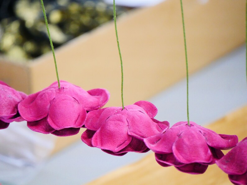 May include: A string of pink paper flowers hanging from green strings. The flowers are made from paper and have a ruffled edge.