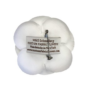 May include: White fabric flower with a pin on the back. The flower has a label that reads "M&S Schmalberg Custom Fabric Flowers Handmade in New York www.customfabricflowers.com"