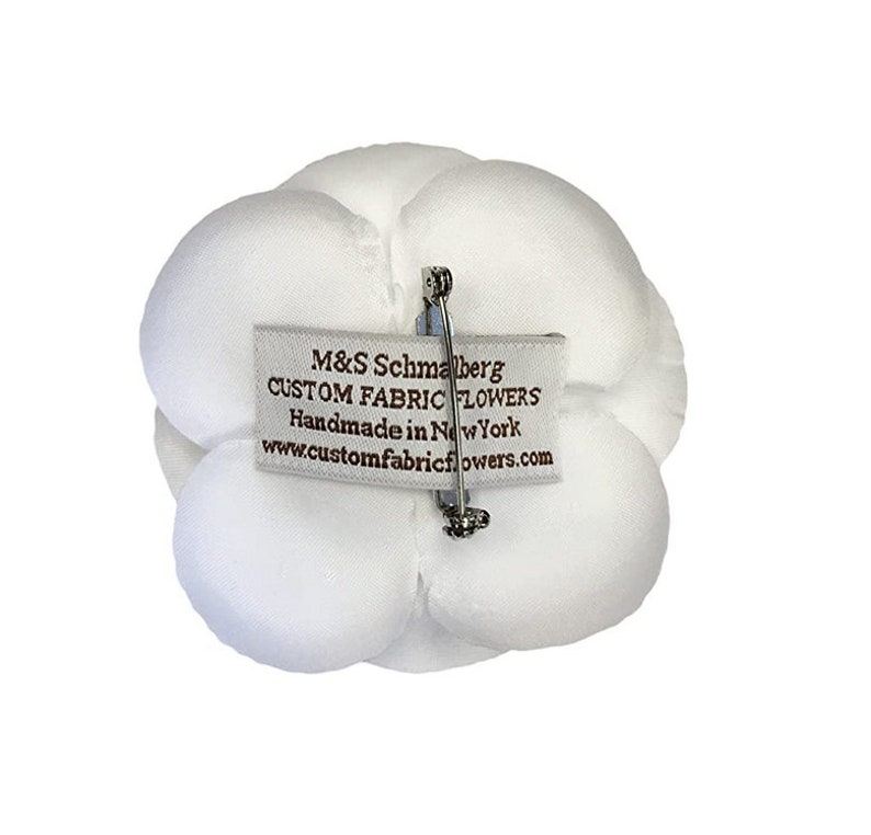 May include: White fabric flower with a pin on the back. The flower has a label that reads "M&S Schmalberg Custom Fabric Flowers Handmade in New York www.customfabricflowers.com"
