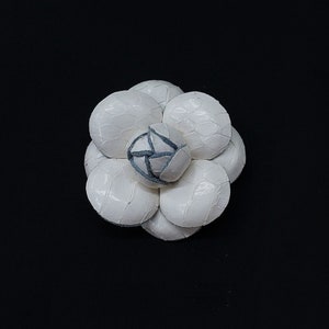 Könnte beinhalten: Eine weiße, blütenförmige Brosche mit strukturierter Oberfläche. Die Brosche besteht aus mehreren abgerundeten Blütenblättern, wobei ein zentrales Blütenblatt ein blau-graues Design aufweist. Der Hintergrund ist einfarbig schwarz.