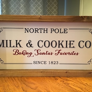 May include: A white sign with black and red lettering in a wooden frame. The sign reads "North Pole Milk & Cookie Co. Baking Santa's Favorites Since 1823".